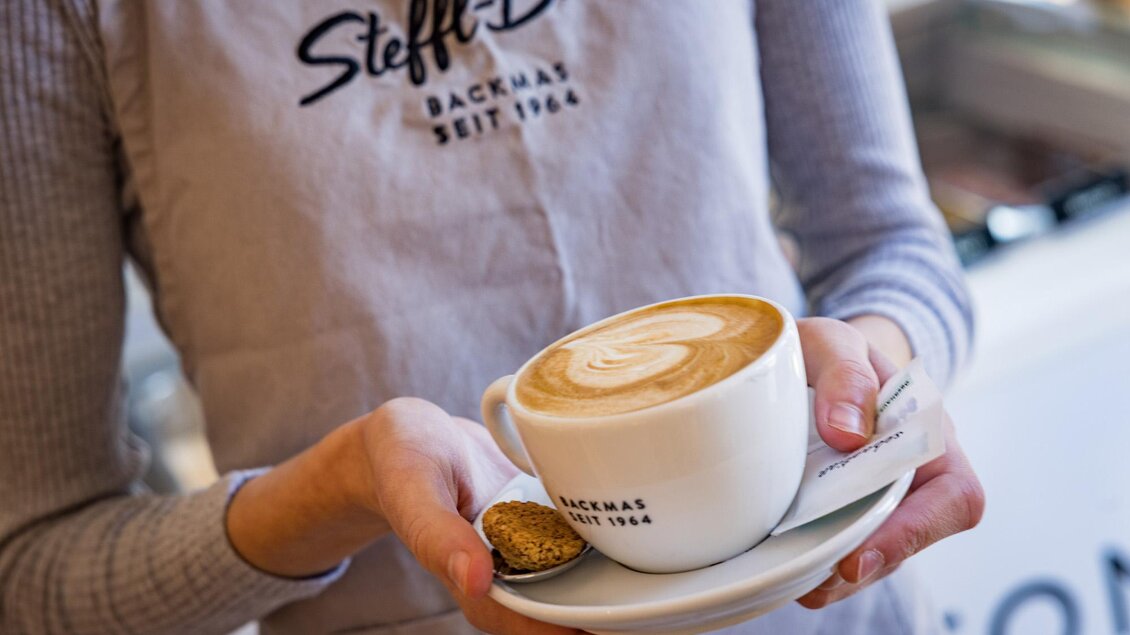 Eine Tasse Kaffee mit einem schönen Milchschaum-Muster wird von einer Person gehalten. Im Hintergrund ist eine helle Café-Umgebung zu sehen. | © Netzwerk Kulinarik wildbild