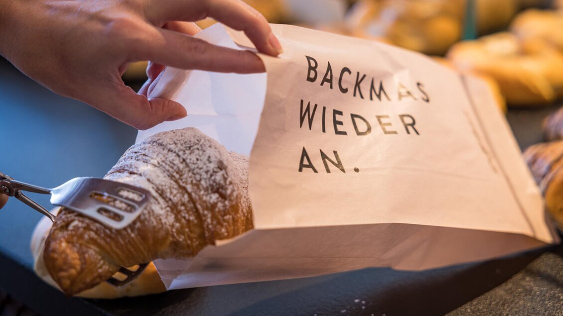 Ein frisches Croissant wird aus einer Tüte mit der Aufschrift „BACKMAS WIEDER AN“ entnommen. Im Hintergrund sind weitere Backwaren sichtbar. | © Netzwerk Kulinarik wildbild