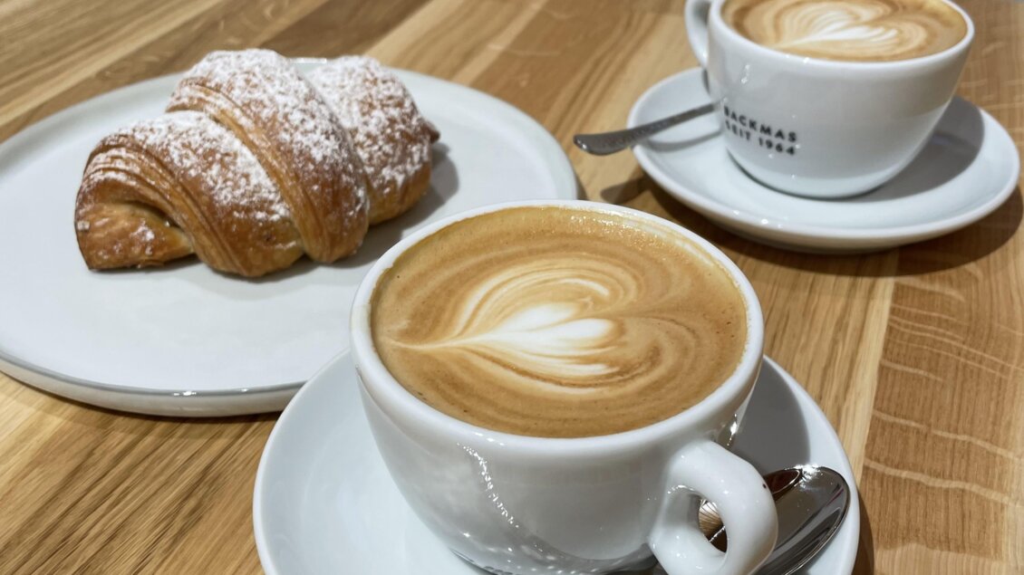 Zwei Tassen Kaffee auf einem Holztisch, begleitet von einem Croissant. Die Tassen sind mit einem hübschen Latte Art-Muster dekoriert. | © Steffl-Bäck Sebastian Knapp