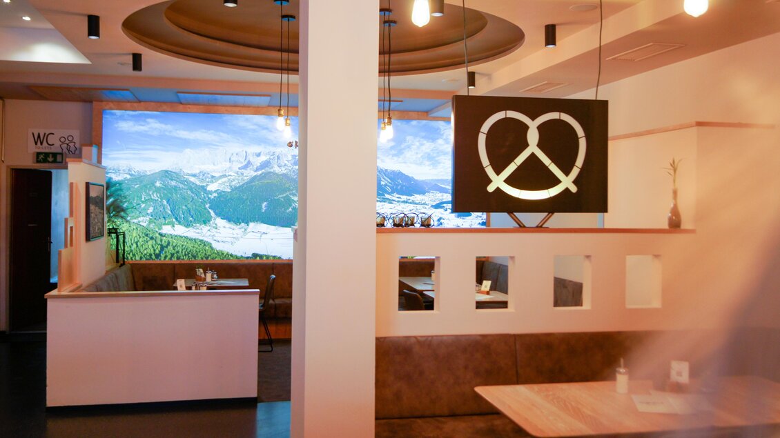 Ein modernes Restaurant mit einer gemütlichen Sitzgelegenheit und einer großen Wandprojektion von Berglandschaften. Ein hängendes Schild in Form einer Brezel ergänzt das einladende Ambiente. | © Steffl-Bäck Sebastian Knapp