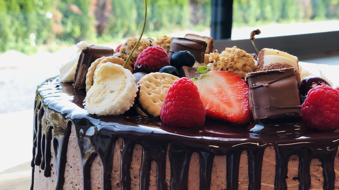 Eine köstliche Schokoladentorte mit frischen Früchten und dekorativen Süßigkeiten. Der Hintergrund zeigt eine grüne Landschaft und Fensterlicht. | © Steffl-Bäck Sebastian Knapp