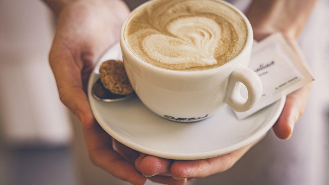 Eine Tasse Kaffee mit kunstvollem Latte Art in den Händen. Auf dem Unterteller liegt ein kleiner Keks und ein Zuckersäckchen. | © AMA Genussregion _wildbild.at