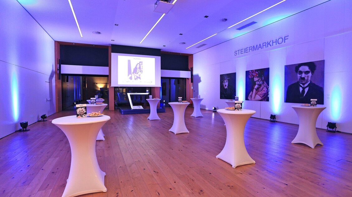 Ein eleganter Veranstaltungsraum mit runden Stehtischen und blauem Licht. An der Wand sind Kunstwerke und ein Projektor zu sehen. | © Steiermarkhof - Foto Pachernegg