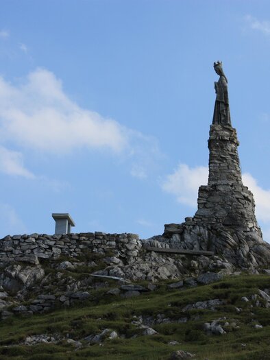 A statue on a high rock with a clear sky in the background. The landscape consists of rocky terrain and gentle hills. | © Willi Fuchs