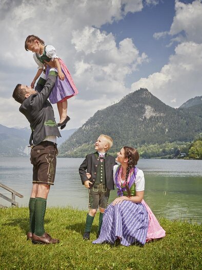 Eine Familie in traditioneller Tracht steht am Ufer eines Sees mit Bergen im Hintergrund. Ein Erwachsener hebt ein Kind in die Luft, während zwei weitere Kinder zuschauen. | © photoInStyle, Birgit Steinberger