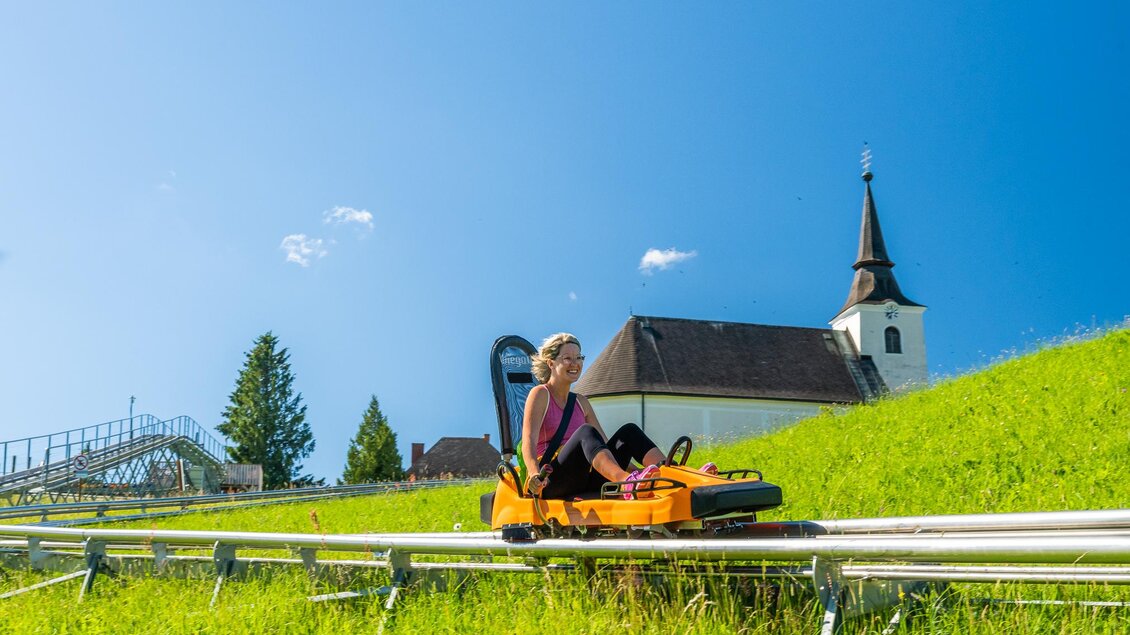 Eine Frau fährt auf einer Sommerrodelbahn in einem grünen Tal. Im Hintergrund ist eine Kirche und ein klarer blauer Himmel zu sehen. | © Region Graz - Mias Photoart