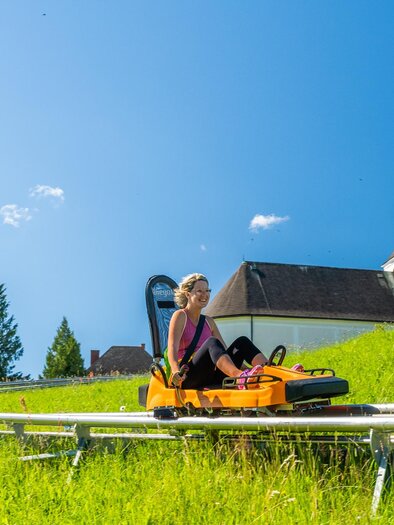 A woman is riding on a summer toboggan run in a green valley. In the background, a church and a clear blue sky can be seen. | © Region Graz - Mias Photoart