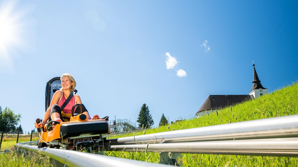 Eine fahrende Person auf einer Sommerrodelbahn in einer grünen Landschaft. Im Hintergrund ist eine Kirche zu sehen, und der Himmel ist klar und sonnig. | © Region Graz - Mias Photoart