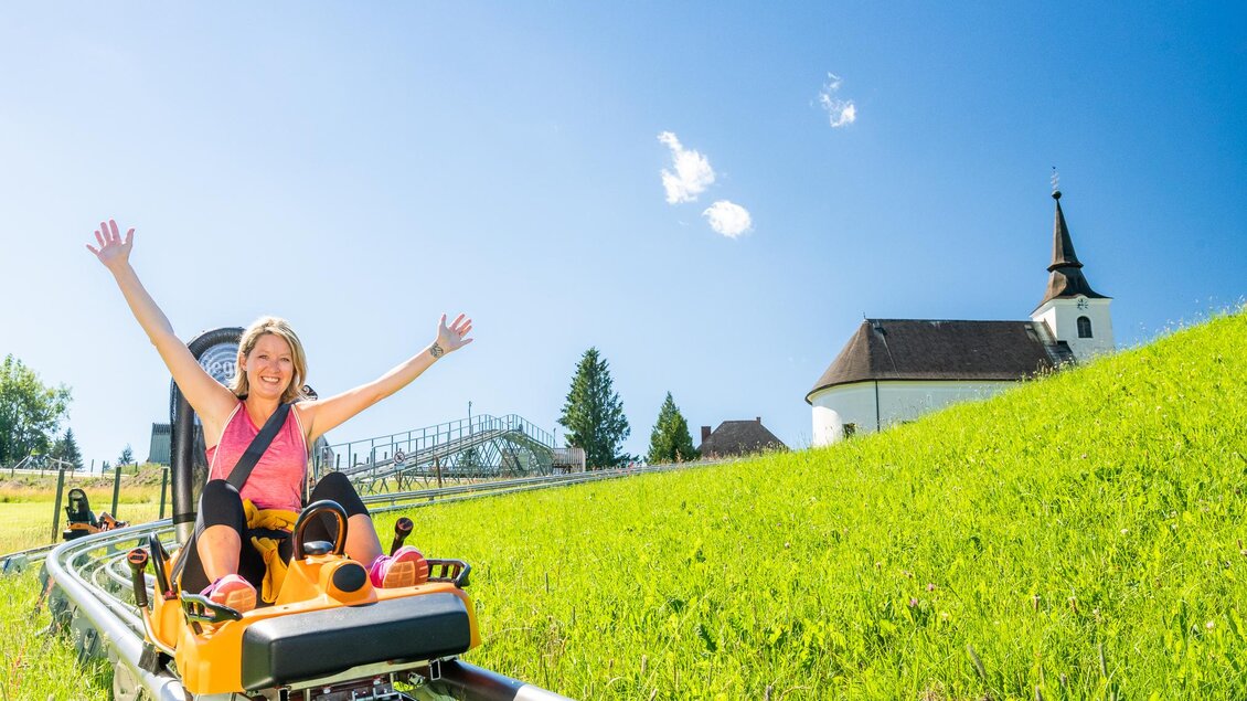 Eine Frau fährt fröhlich auf einer Rodelbahn den Hügel hinunter. Im Hintergrund sind eine grüne Wiese und ein Kirchturm zu sehen. | © Region Graz - Mias Photoart