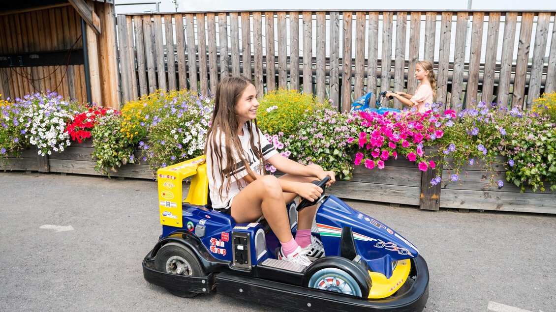 Ein Mädchen fährt in einem Karts und lächelt. Im Hintergrund stehen bunte Blumen und ein weiteres Kind spielt. | © Diesel