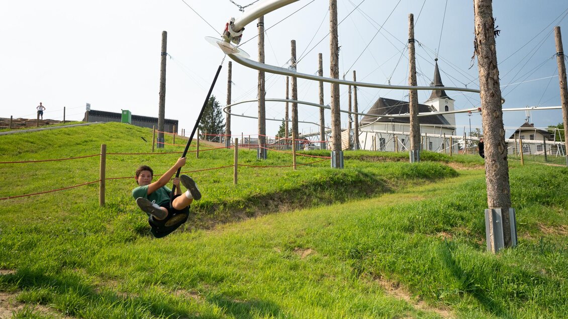 Ein Junge schwingt auf einer Seilbahn über eine grüne Wiese. Im Hintergrund sind Bäume und eine Kirche zu sehen. | © Diesel