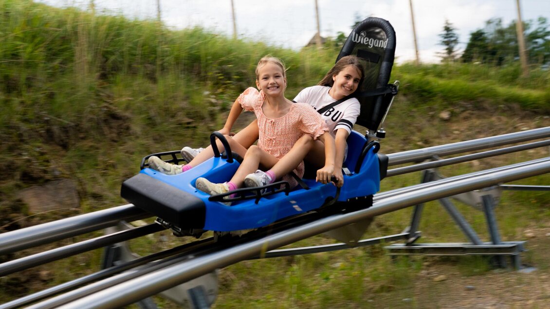 Zwei Mädchen fahren auf einer Sommerrodelbahn. Sie lächeln und haben Spaß während der Fahrt durch die grüne Landschaft. | © Diesel