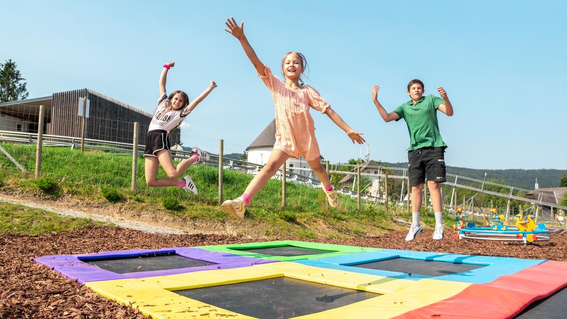 Drei Kinder springen fröhlich auf bunten Trampolinen im Freien. Im Hintergrund sind Wiesen und Gebäude sichtbar. | © Diesel
