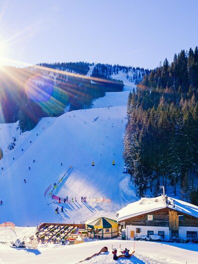 A snow-covered landscape with skiers on the slopes. In the foreground are huts and trees, while the sun shines brightly over the mountains. | © Roland Schoblocher