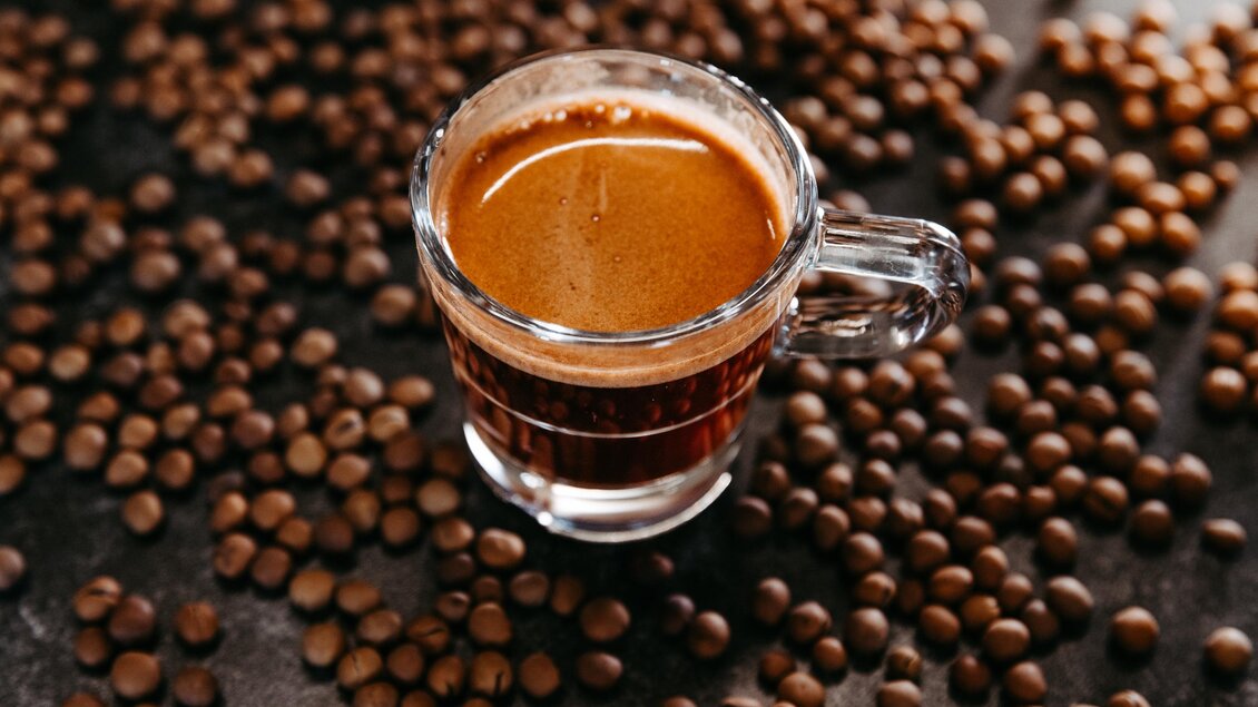 Ein Glas frischer Espresso steht auf einer Unterlage aus Kaffeebohnen. Der Kaffee hat eine reichhaltige, dunkle Farbe und eine cremige Schicht oben. | © Region Graz - Mias Photoart