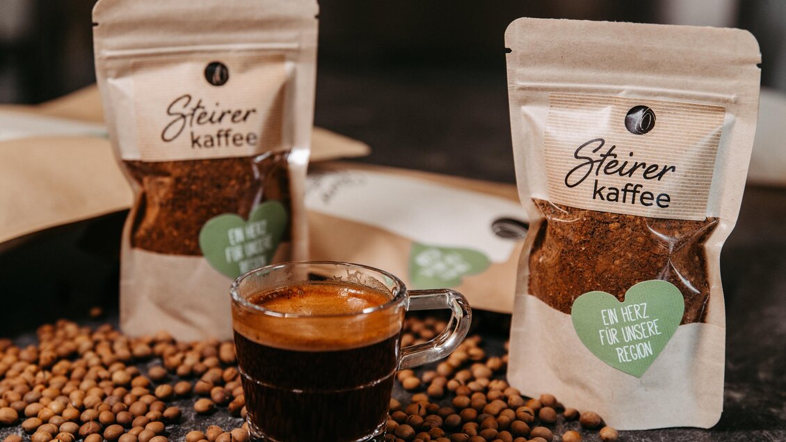 Ein Glas frischer Kaffee steht vor zwei Verpackungen von Steirer Kaffee. Kaffeebohnen liegen verstreut auf der Tischfläche. | © Region Graz - Mias Photoart