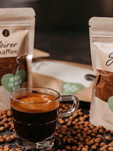 Ein Glas frischer Kaffee steht vor zwei Verpackungen von Steirer Kaffee. Kaffeebohnen liegen verstreut auf der Tischfläche. | © Region Graz - Mias Photoart