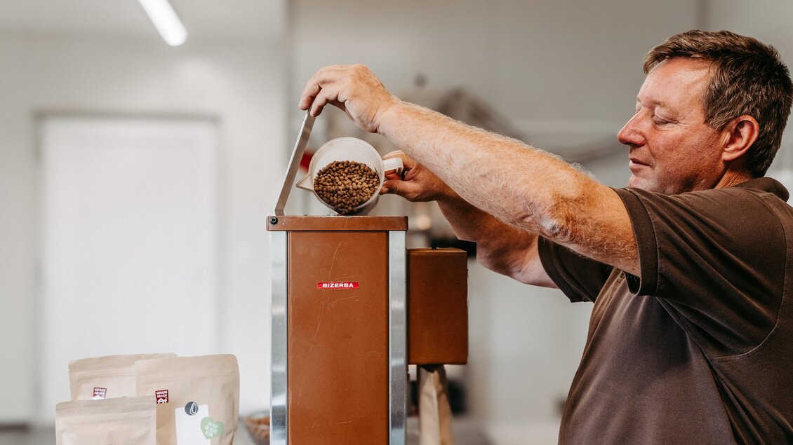 Ein Mann füllt Kaffeebohnen in eine Maschine. Im Hintergrund sind einige Kaffeepackungen sichtbar. | © Region Graz - Mias Photoart