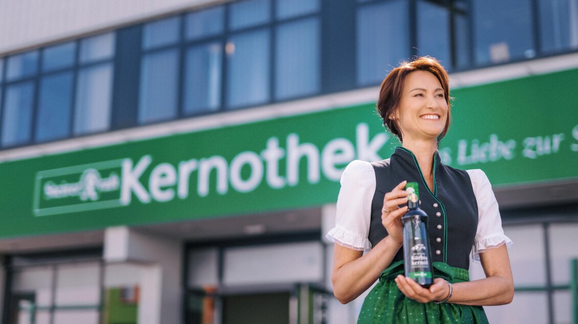Eine lächelnde Frau in traditoneller Tracht hält eine Flasche in der Hand. Im Hintergrund ist das Geschäftsschild "Kernothek" sichtbar. | © Steirerkraft