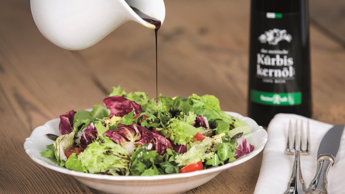Ein frischer Salat mit buntem Gemüse, über den ein Dressing gegossen wird. Daneben steht eine Flasche Kürbiskernöl. | © Steirerkraft