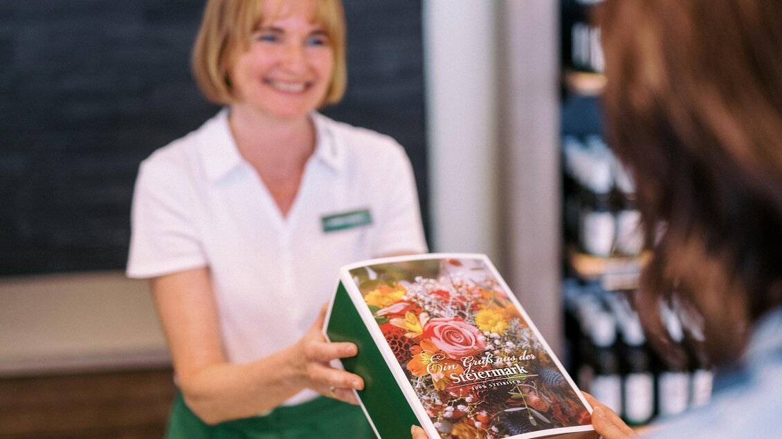 Eine freundliche Verkäuferin übergibt eine Box mit Blumen oder Geschenken an eine Kundin. Im Hintergrund sind Weinflaschen und ein Holzschild zu sehen. | © Steirerkraft
