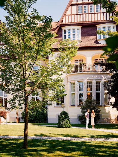A beautiful manor house surrounded by a well-maintained garden. Two people are standing in front of the facade, enjoying the sunny atmosphere. | © Tauroa GmbH