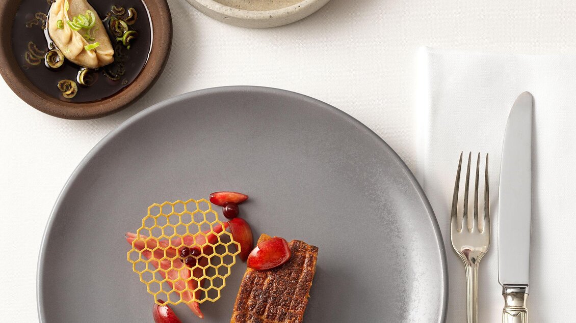 Ein stilvolles Gericht auf einem grauen Teller mit Honigwaben und einer roten Sauce. Dazu ein Glas Rotwein und kleine Beilagen in einer Schale. | © Steirerschlössl