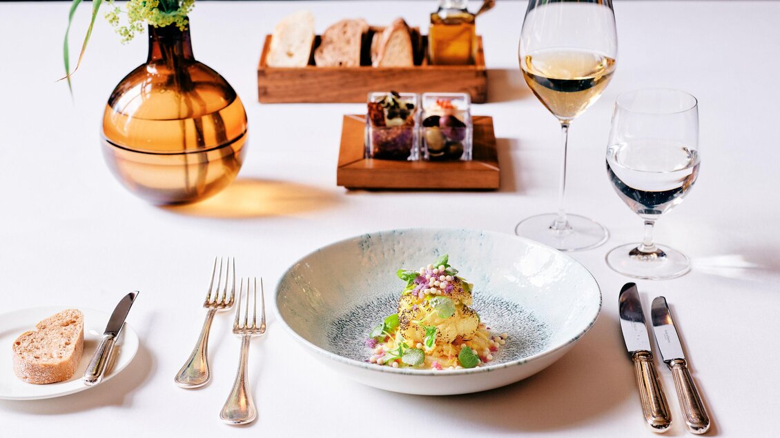 Ein elegant gedeckter Tisch mit einem köstlichen Gericht in einer Schüssel. Neben dem Essen stehen Wein und Wasser, sowie frisches Brot in einer Holzschale. | © Steirerschlössl