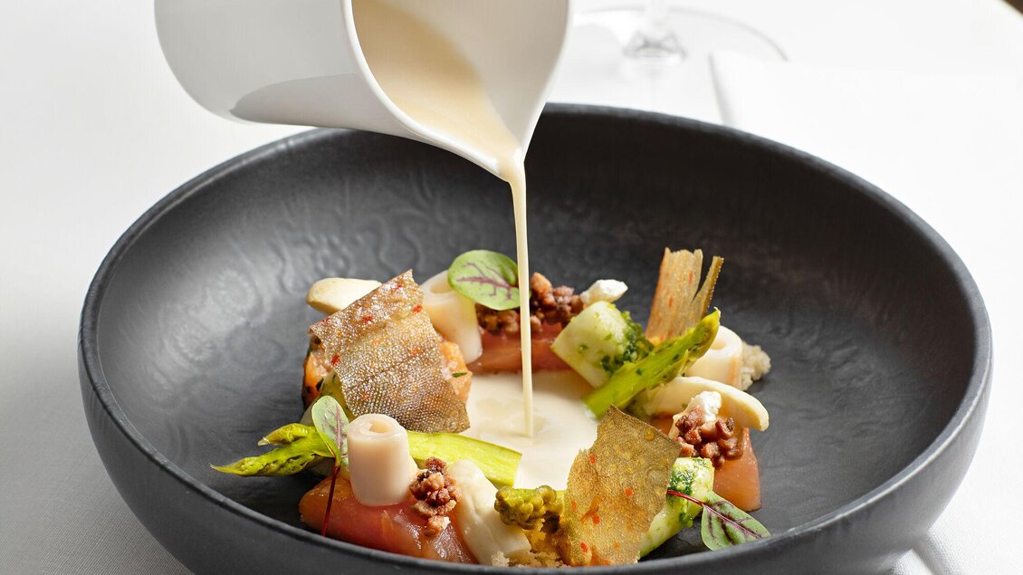 Ein kunstvoll angerichteter Teller mit geröstetem Gemüse und einer cremigen Sauce. Im Hintergrund ist ein Glas Wein sichtbar. | © Steirerschlössl