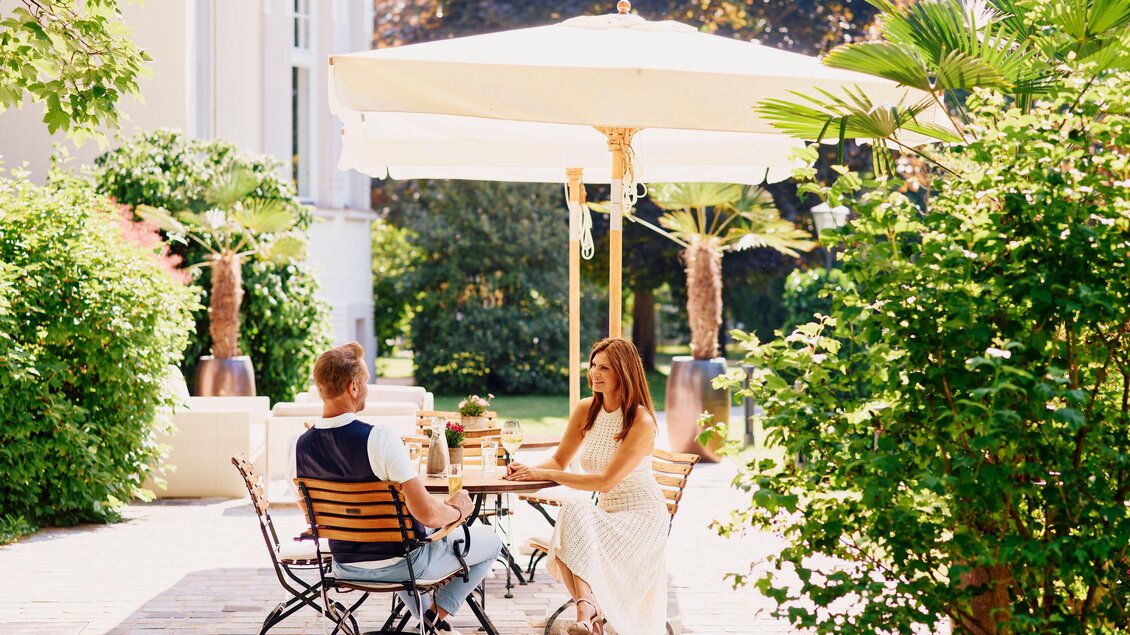 Ein Paar sitzt entspannt unter einem Sonnenschirm in einem schönen Garten. Grüne Pflanzen umgeben sie und schaffen eine gemütliche Atmosphäre. | © Steirerschlössl
