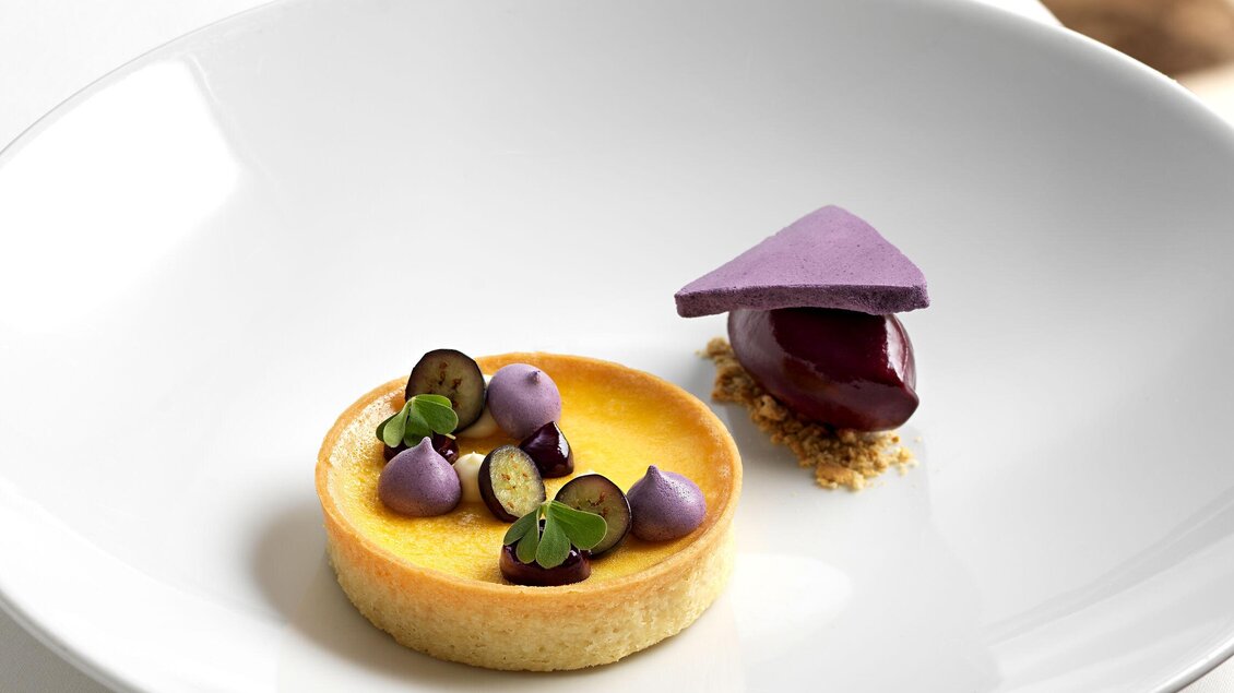 Ein kunstvoll angerichteter Dessertteller mit einer Zitronentorte und lila Dekorationen. Die Torte ist auf einem weißen Teller mit einem eleganten Design platziert. | © Steirerschlössl