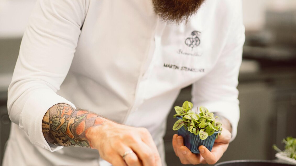 Ein Koch in weißer Kleidung arbeitet an der Präsentation eines Gerichts. Er dekoriert einen Teller mit frischen Kräutern und lächelt dabei. | © Steirerschlössl