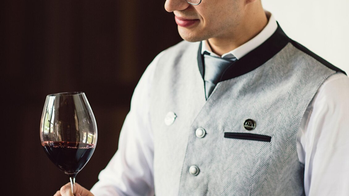 Ein Kellner in einer eleganten Tracht hält ein Weinglas mit Rotwein in der Hand. Er blickt aufmerksam auf den Wein und strahlt Professionalität aus. | © Tauroa GmbH