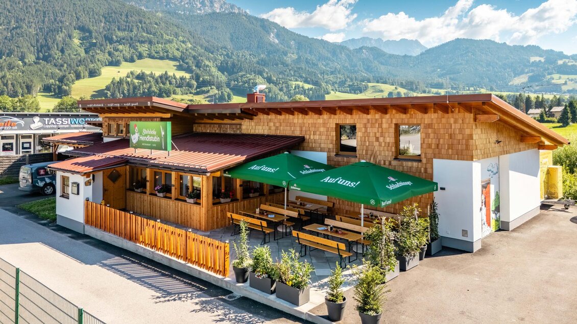 Ein einladendes Restaurant mit traditionellen Holzelementen und grünen Markisen. Im Hintergrund sind malerische Berge und eine grüne Landschaft zu sehen. | © Steirische Hendlstub'n