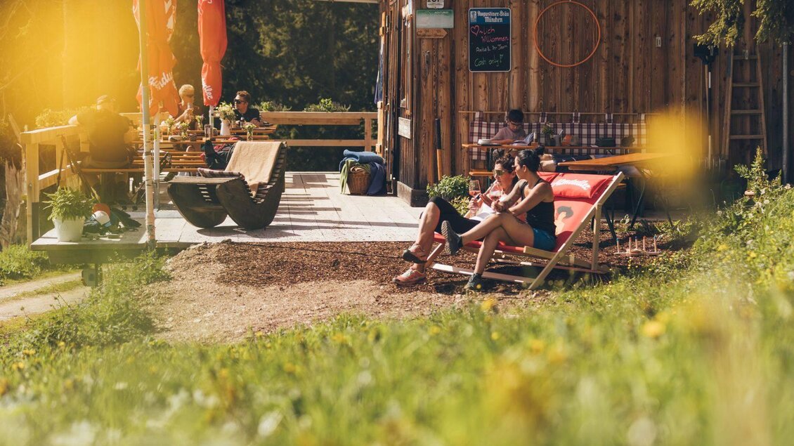 Eine gemütliche Terrasse mit Sonnenliegen und Gästen, die sich entspannen. Im Hintergrund steht ein Holzgebäude, umgeben von einer grünen Wiese. | © Krewenka
