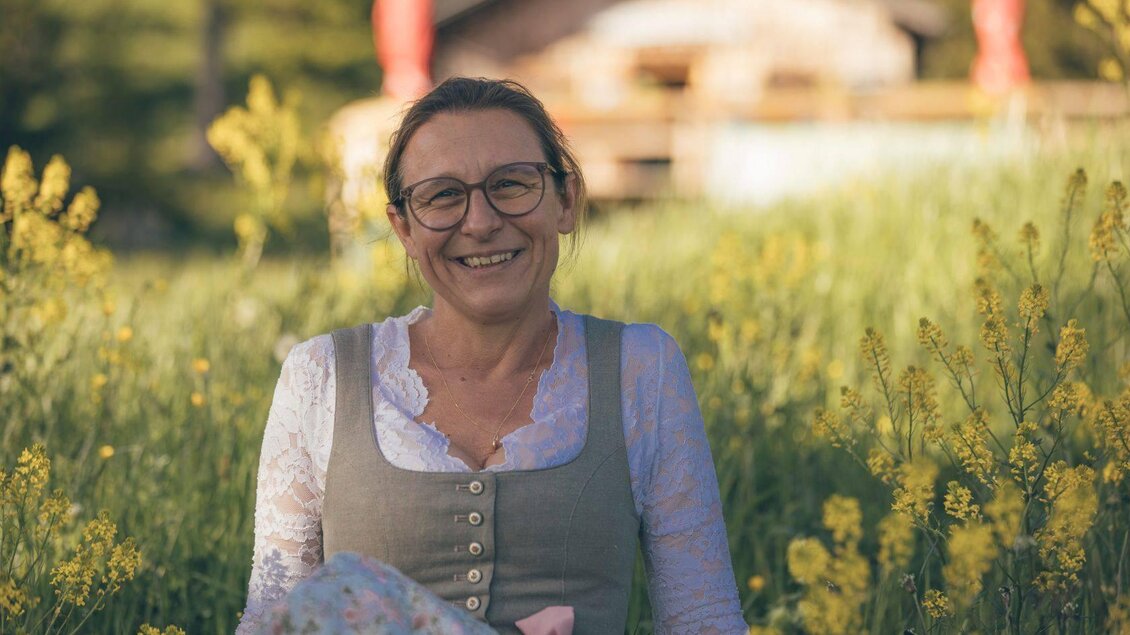 Eine lächelnde Frau sitzt in einem blühenden Feld voller gelber Blumen. Im Hintergrund ist ein Holzhaus und Bäume zu sehen. | © Krewenka