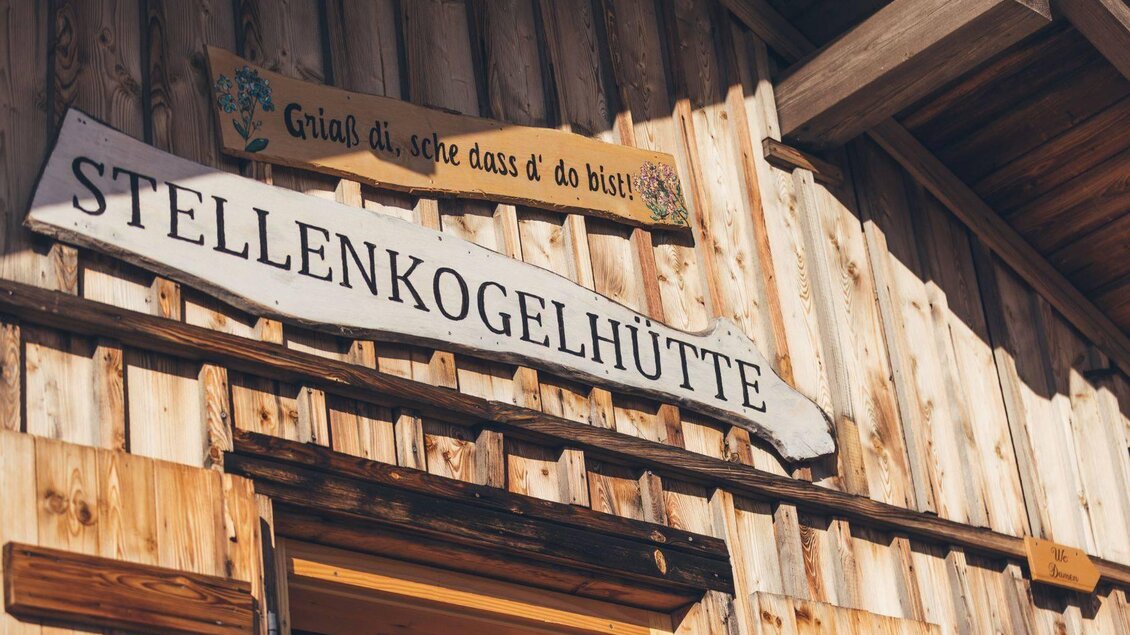 Ein Holzhaus mit dem Schild "Stellenkogelhütte". Oben hängt ein weiteres Schild mit der Begrüßung "Griaß di, schee dass d’ do bist!". | © Krewenka