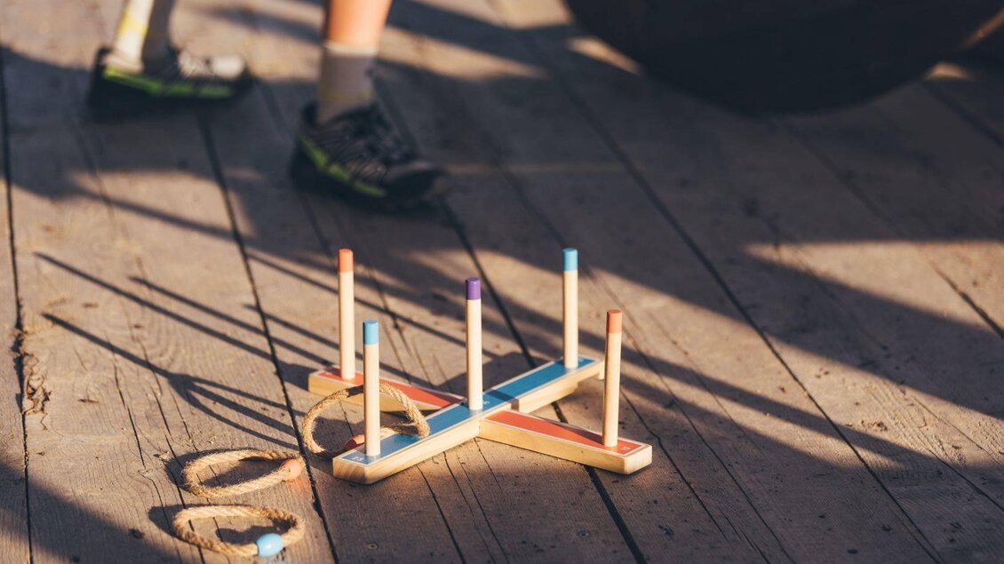 Ein Holzspielzeug mit bunten Stäben und Ringen liegt auf einem Holzboden. Im Hintergrund sind Beine in Sportschuhen sichtbar. | © Krewenka