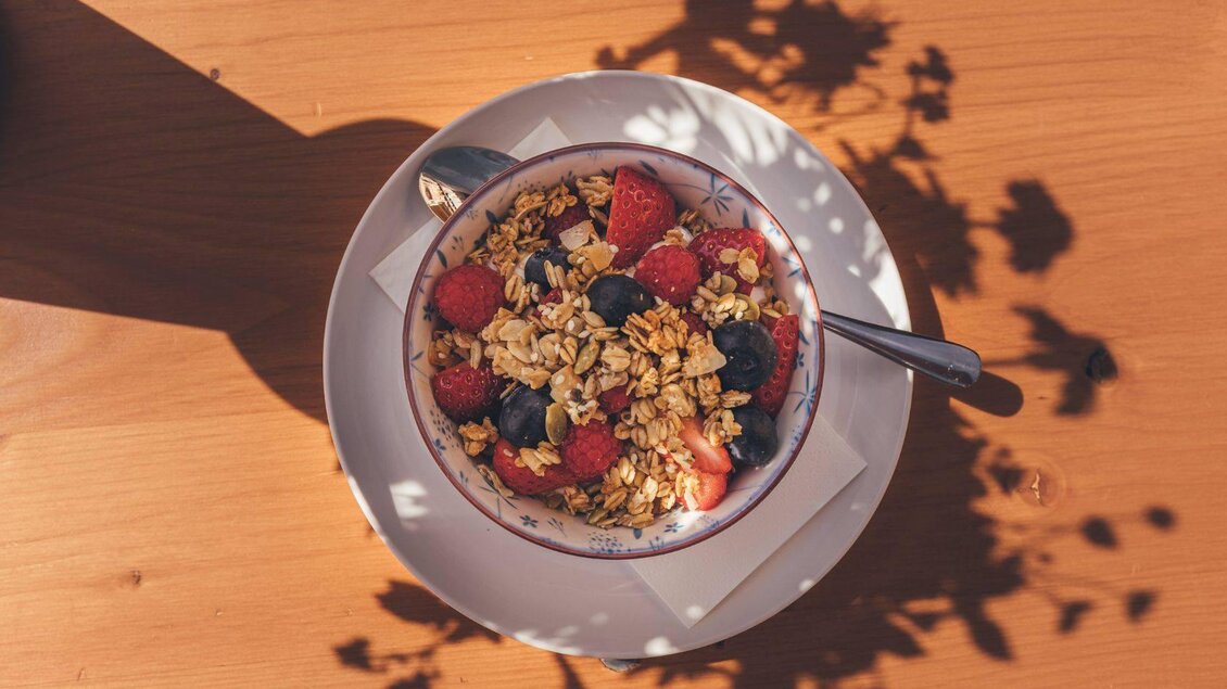 Eine Schüssel Müsli mit frischen Beeren auf einem Holztisch. Der Schatten von Pflanzen zeichnet interessante Muster auf die Oberfläche. | © Krewenka