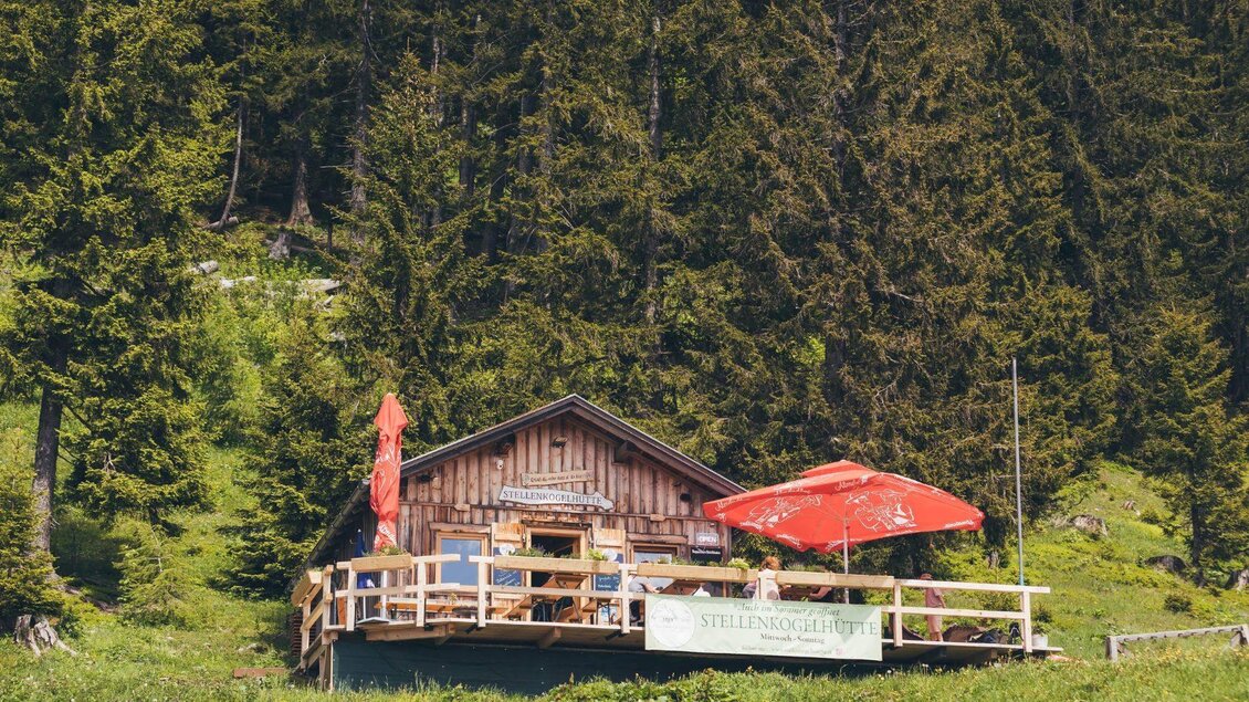 Eine gemütliche Berghütte umgeben von grünen Wäldern. Auf der Terrasse stehen Tische mit roten Sonnenschirmen. | © Krewenka