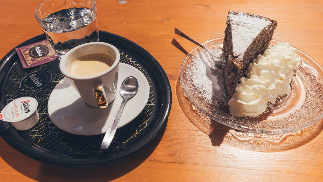 Eine Tasse Kaffee mit einem Stück Schokoladentorte und Sahne auf einem Teller. Nebenbei steht ein Glas Wasser auf einem Tablett. | © Krewenka
