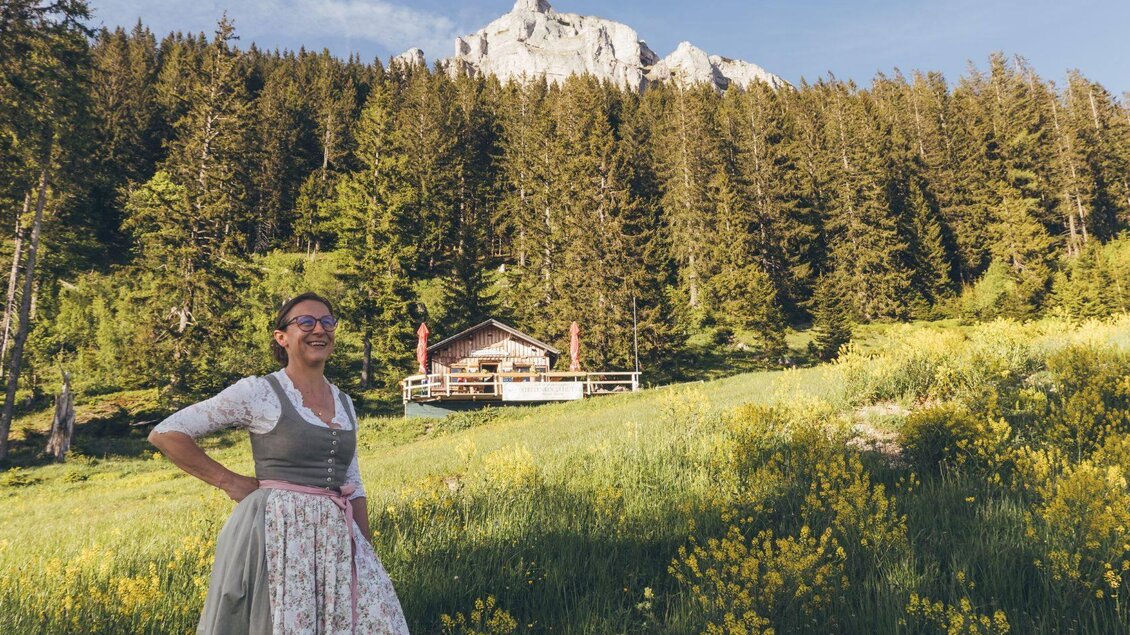 Eine lächelnde Frau in traditioneller Tracht steht auf einer blühenden Wiese. Im Hintergrund sind Bäume und ein Berg zu sehen, sowie eine Hütte. | © Krewenka