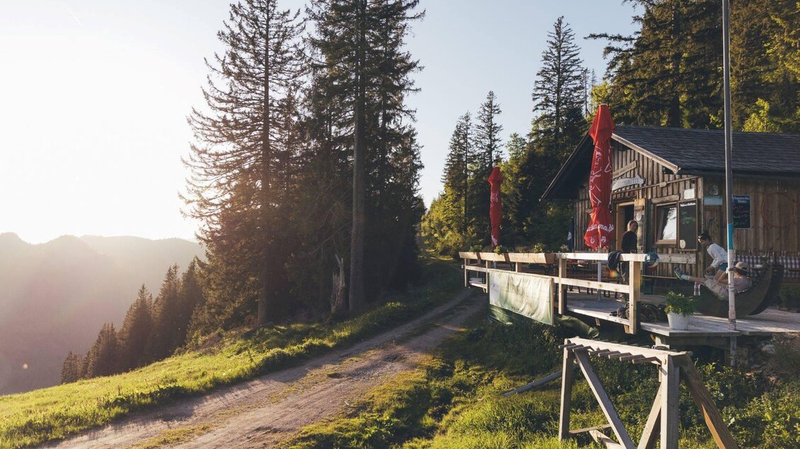 Eine Berghütte am Weg mit Blick auf die Berge. Im Hintergrund stehen hohe Bäume und der Sonnenuntergang sorgt für eine warme Atmosphäre. | © Krewenka