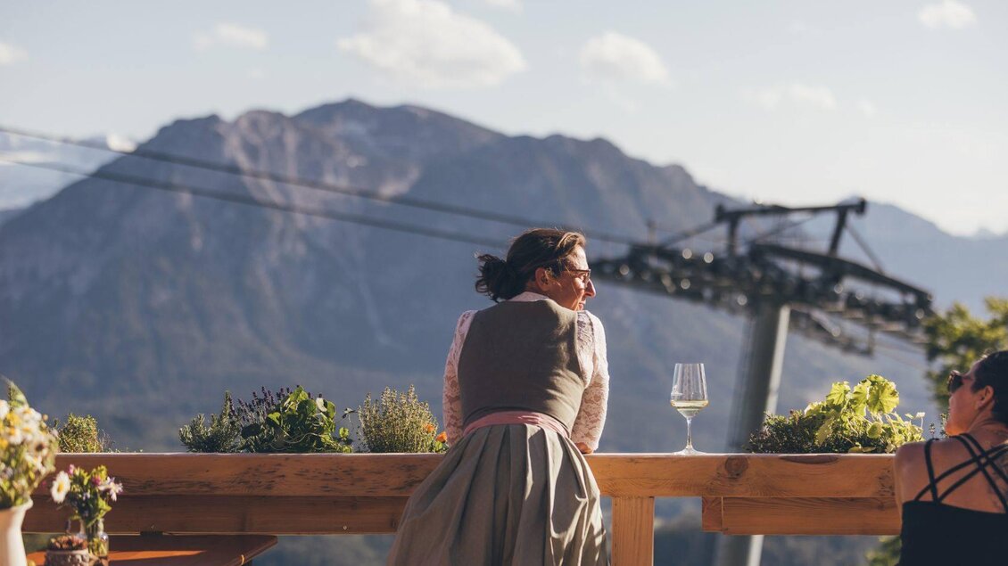 Eine Frau in traditioneller Tracht sitzt an einem Holzgeländer und blickt auf die Berge. Im Hintergrund sind Bergbahnen und grüne Pflanzen zu sehen. | © Krewenka