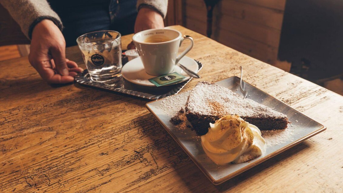 Ein Teller mit Schokoladenkuchen und einer Portion Sahne steht auf einem Holztisch. Daneben befindet sich eine Tasse Kaffee und ein Glas Wasser. | © Krewenka