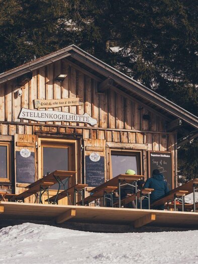 Eine gemütliche Skihütte aus Holz, umgeben von Schnee. Tische und Stühle stehen auf der Terrasse, ideal für eine Pause nach dem Skifahren. | ©  Krewenka