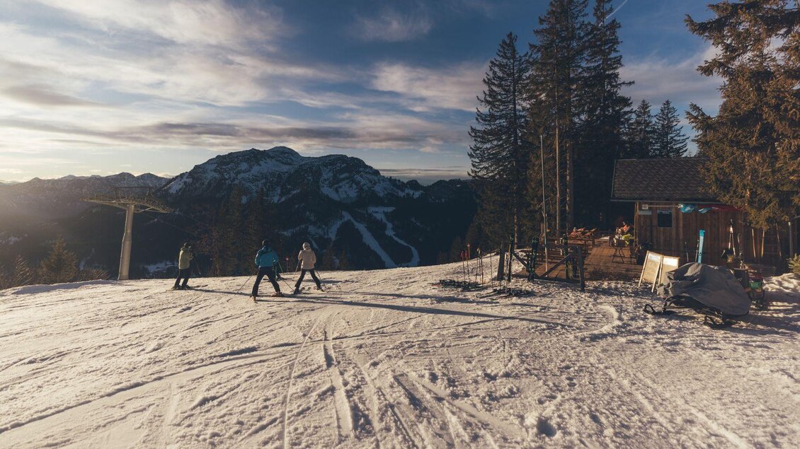 Eine schneebedeckte Piste mit Skifahrern und umliegenden Bergen. Im Hintergrund sind Bäume und eine kleine Hütte zu sehen. | © Krewenka