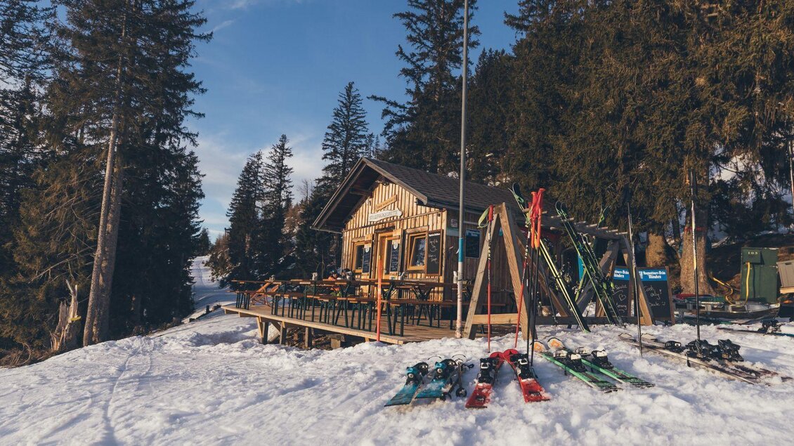 Eine gemütliche Skihütte in der verschneiten Landschaft. Umgeben von hohen Bäumen stehen Skiausrüstungen bereit. | © Krewenka