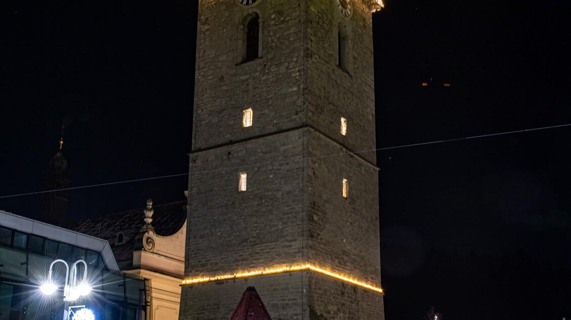 Ein beleuchteter Glockenturm in der Nacht, umgeben von festlicher Dekoration. Der Platz ist weihnachtlich geschmückt mit Lichtern und einem Weihnachtsbaum. | © Planetarium Judenburg