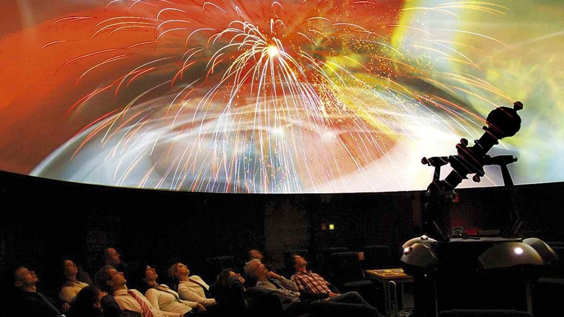 Eine Gruppe von Menschen sitzt in einem Planetarium und schaut auf eine faszinierende Lichtshow am Kuppelhimmel. Der Raum ist mit einem Teleskop ausgestattet und die Farben der Animation wirken lebhaft und spektakulär. | © Horst Kaiser